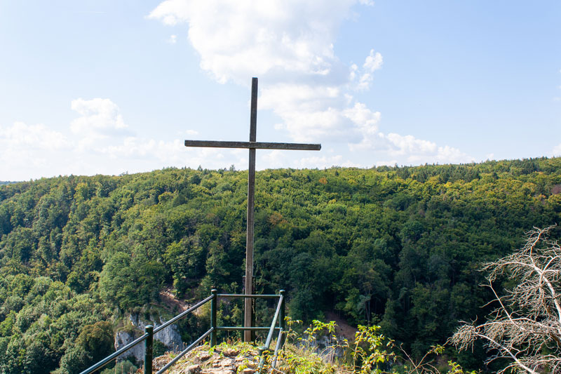 Holzkreuz auf einem Aussichtspunkt über bewaldeter Landschaft mit Blick ins Tal.